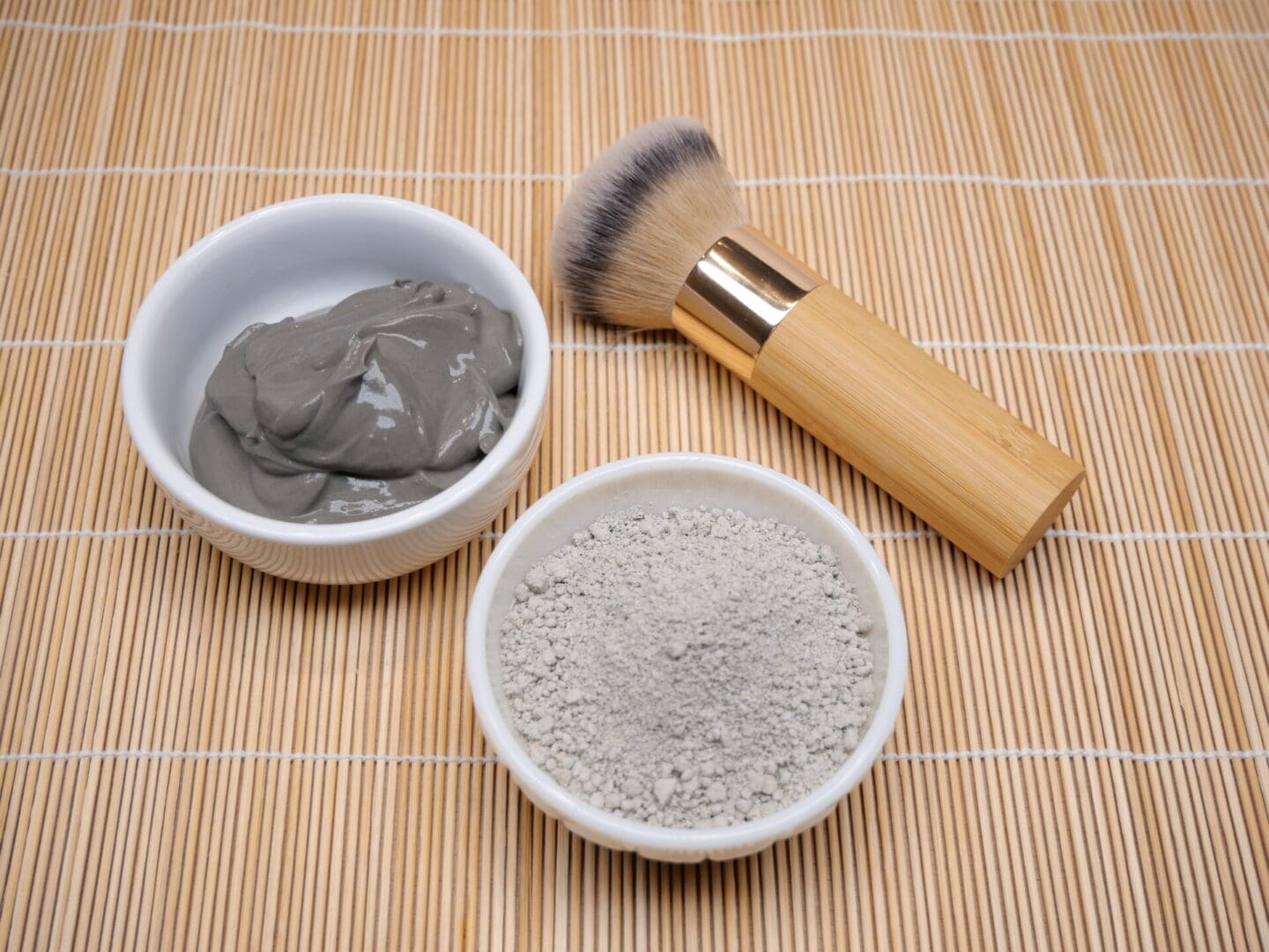 Two bowls of French Green Clay Face Mask Powder Clarifying & Pore Minimizer and a brush on a bamboo mat.