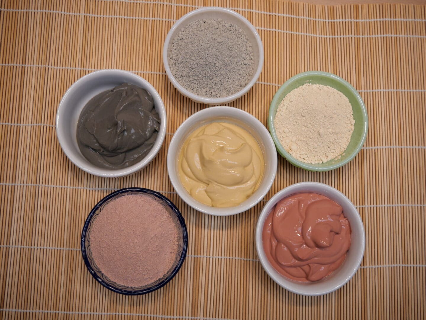 Five bowls of different colored powders, including a French Pink Clay Face Mask Powder for Dry, Sensitive Skin, on a bamboo mat.