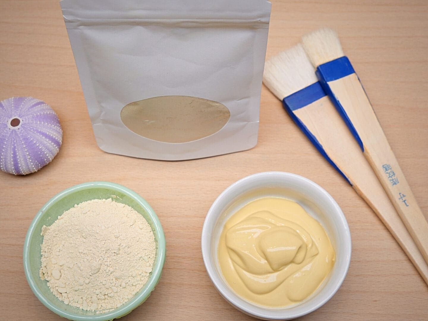 A bowl of Brazilian Yellow Clay Antioxidant Face Mask Powder, brushes, and a bag of powder.
